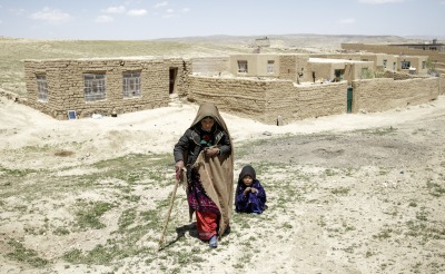 A mother walks away from her home with the help of a cane. Her daughter crouches behind her.
