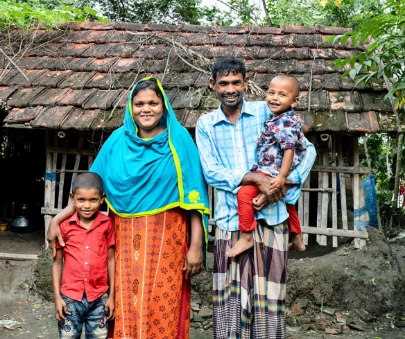 A mother and a father stand smiling with their two sons outside of their home in Bangladesh.