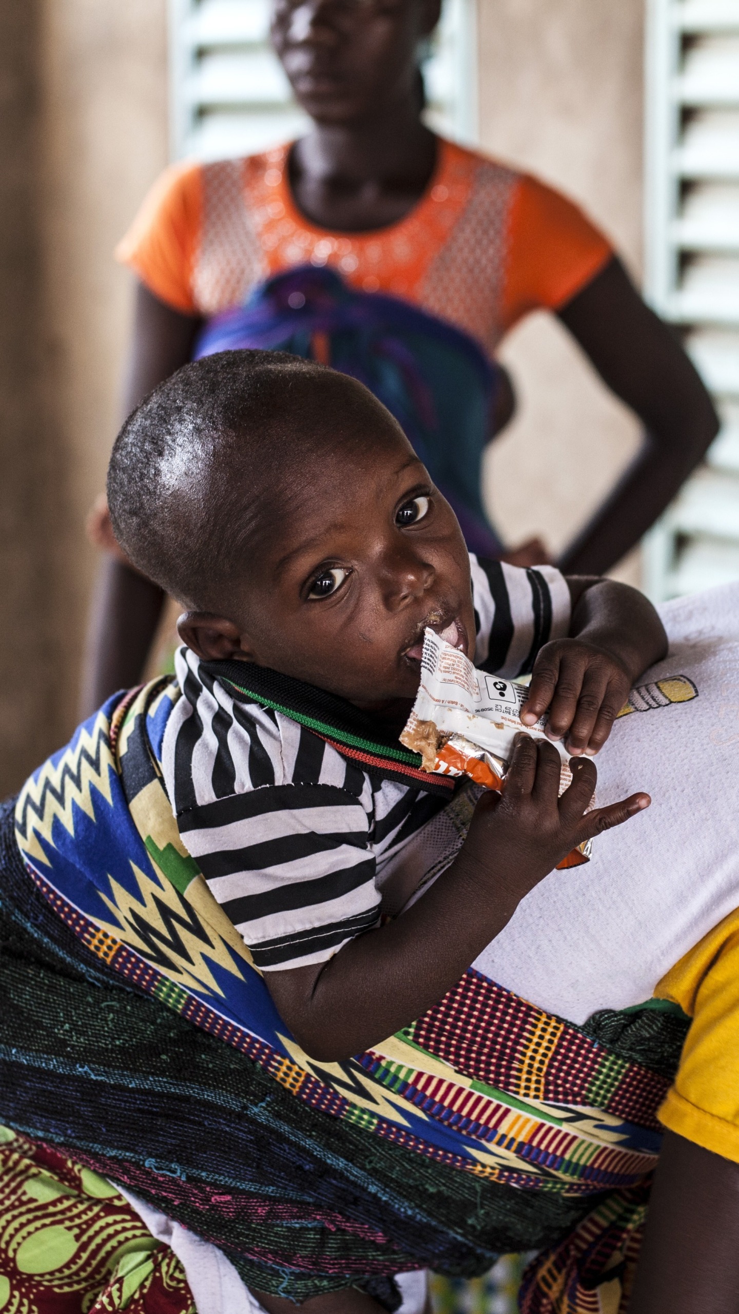 A young boy, on his mother's back, eats Plumpy'Nut to recover from malnutrition.