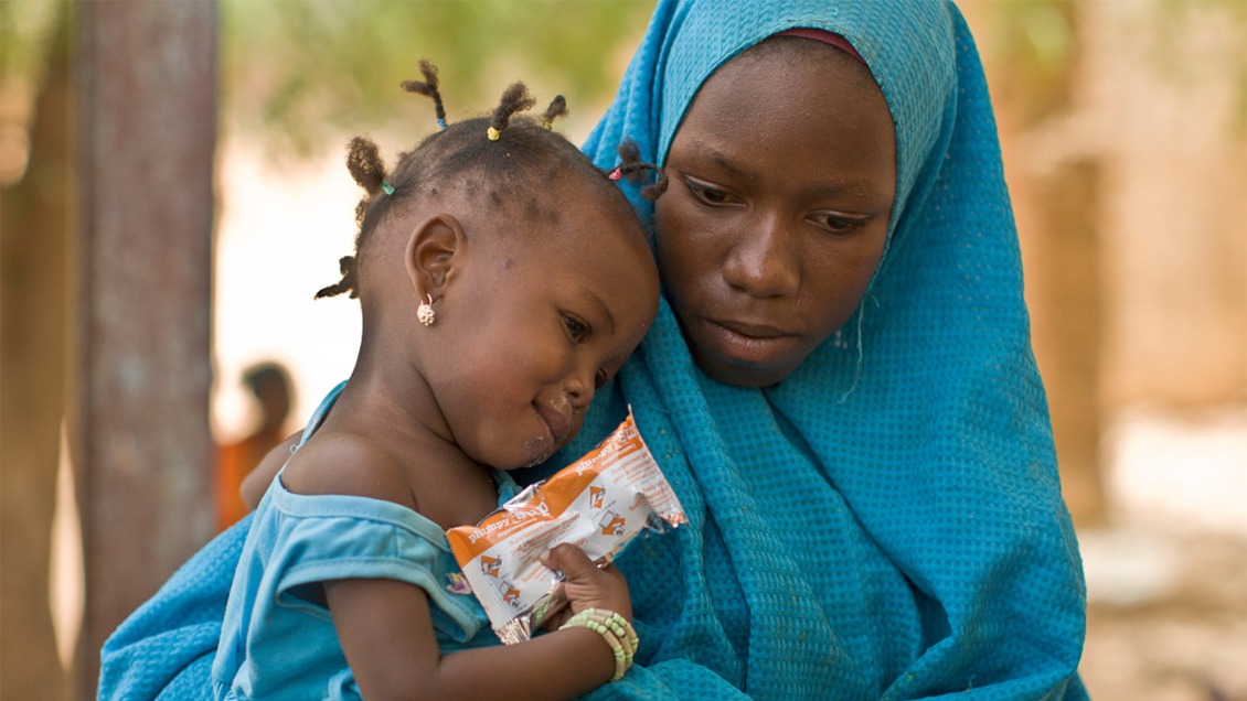A woman holds her little girl who clutches a packet of RUTF.