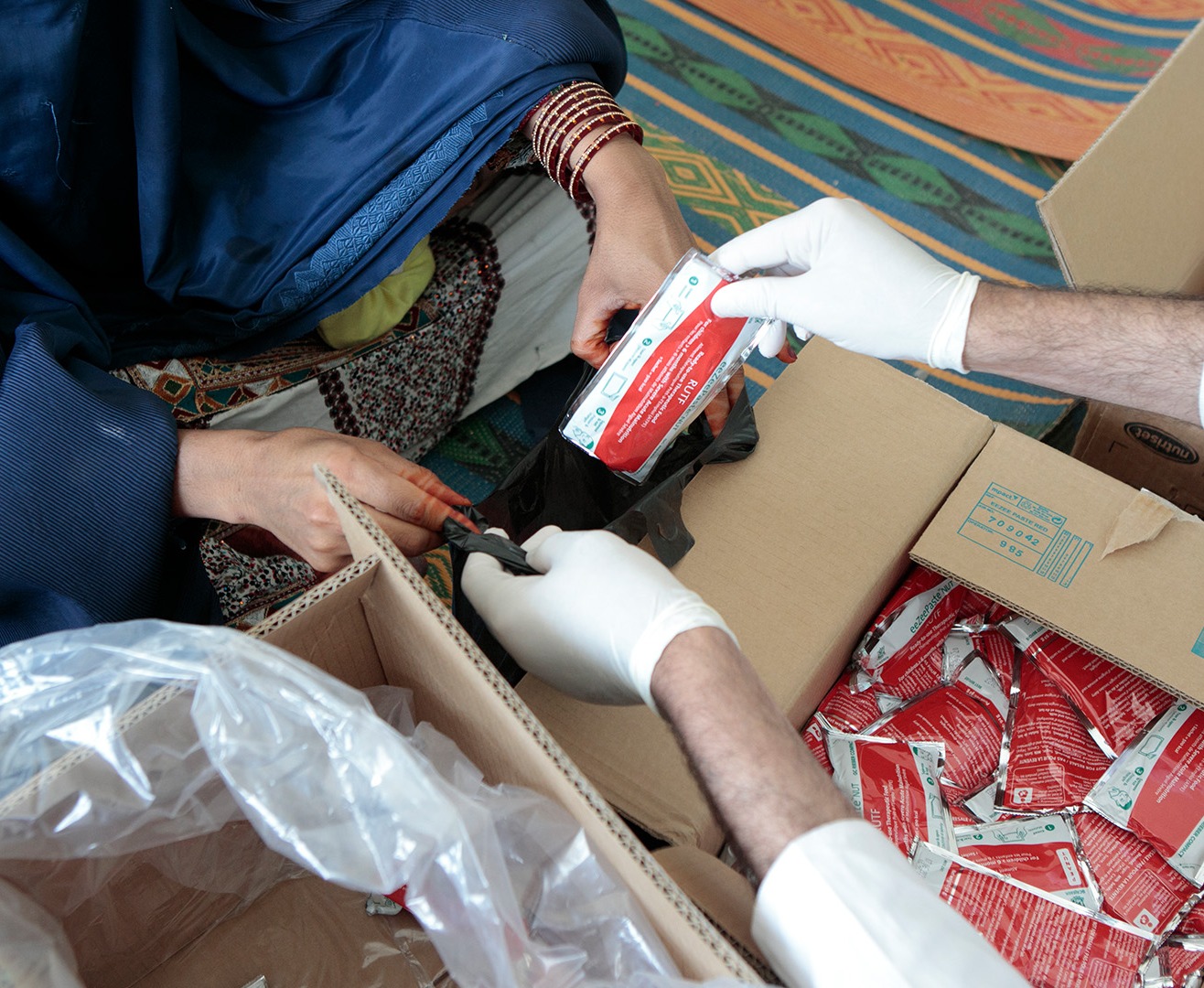 Gloved hands unpacking RUTF packages from a cardboard box and handing one to a woman.