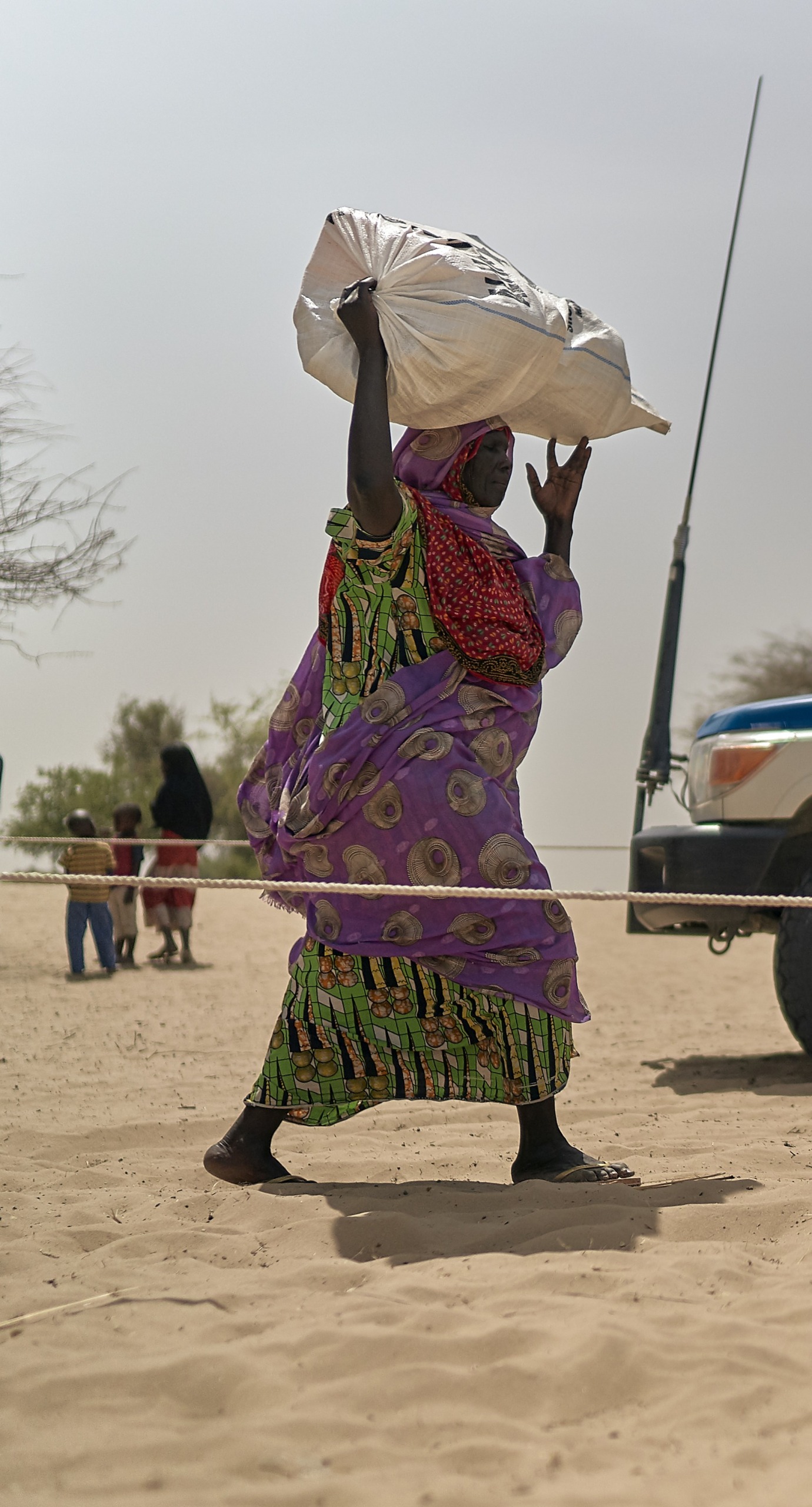 A woman walks through dry, sandy land with a large bag above her head. An Action Against Hunger truck is in the background.